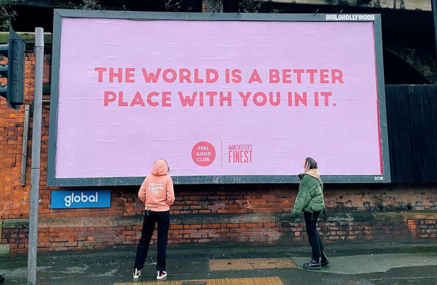 Two women standing in front of a billboard which reads 'The World is a better place with you in it